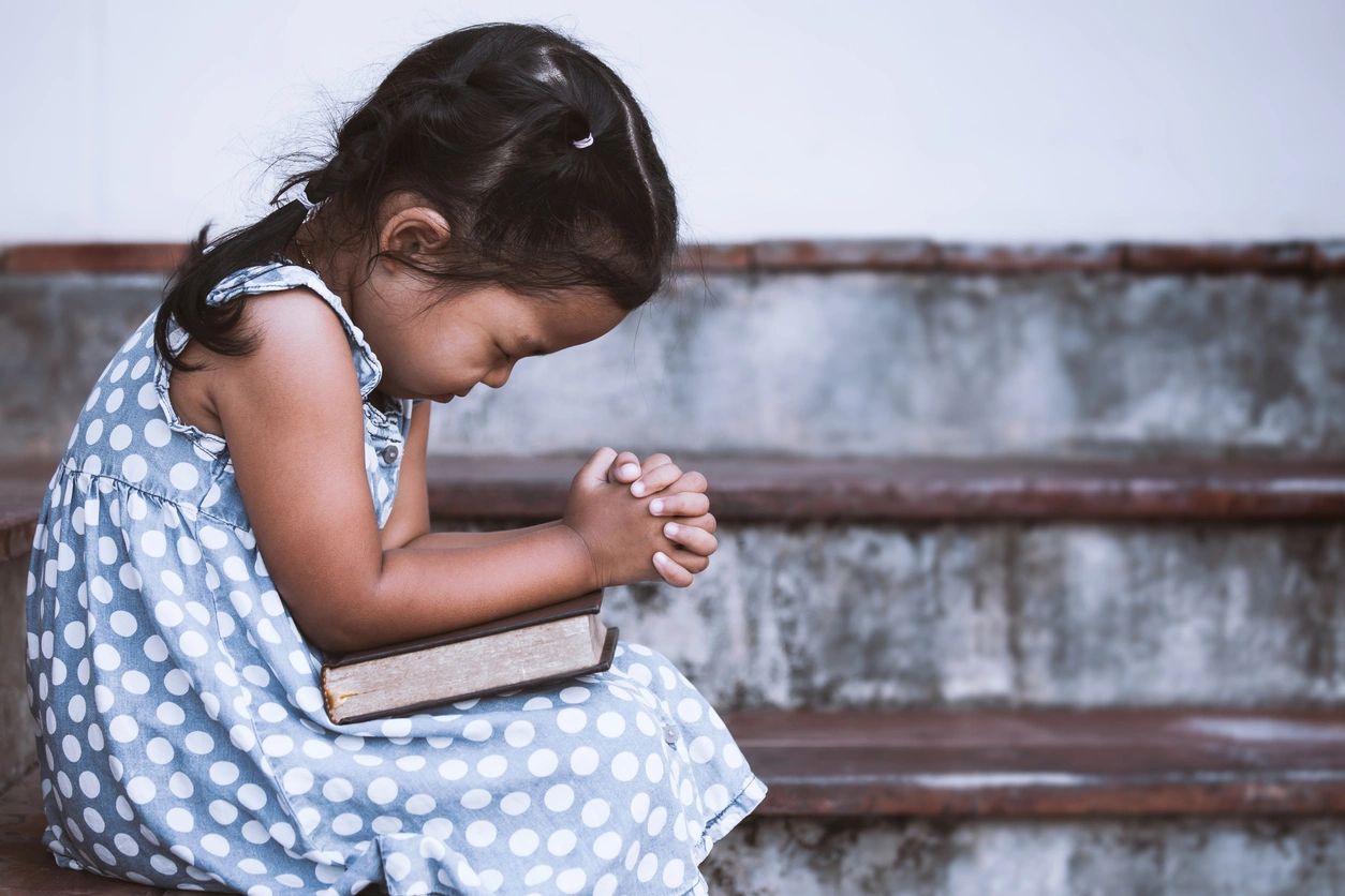 Young girl in a polka-dot dress praying on a staircase with a book beside her.