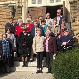 Group of elderly adults smiling on church steps, a mix of standing and sitting, with a historical building backdrop.