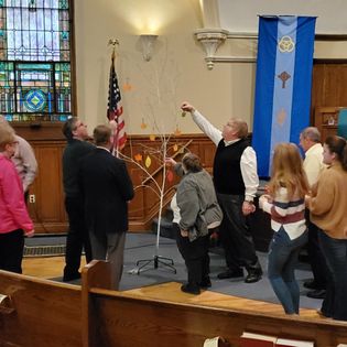 A group of people in a church hall, one man pointing upwards, gathering around a small tree, with the u.s. flag and a banner in the background.