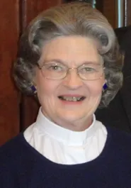 An older woman smiling at the camera, wearing glasses, a blue sweater, and a white collared shirt.